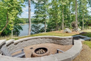 A stone fire pit surrounded by a curved stone bench, overlooking a lake with trees in the background. - Roxboro Lake Family Retreat and Spa (Prospect Hill)