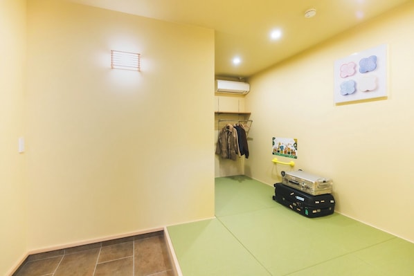 A room with a green floor mat, a wall-mounted air conditioner, a clothes rack with jackets, and a suitcase on the floor.