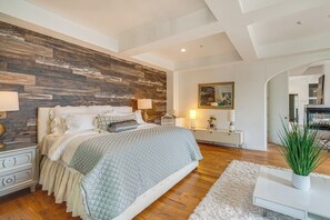 Ein Schlafzimmer mit Holz-Kopfteil, einem Bett mit vielen Kissen, Nachttischen mit Lampen, einem weißen Teppich und einer Pflanze auf einem Beistelltisch.