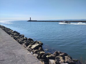 A coastal scene with a lighthouse, a concrete walkway, and a boat creating waves. - Calm sea. 50 m² apartment (Agde)
