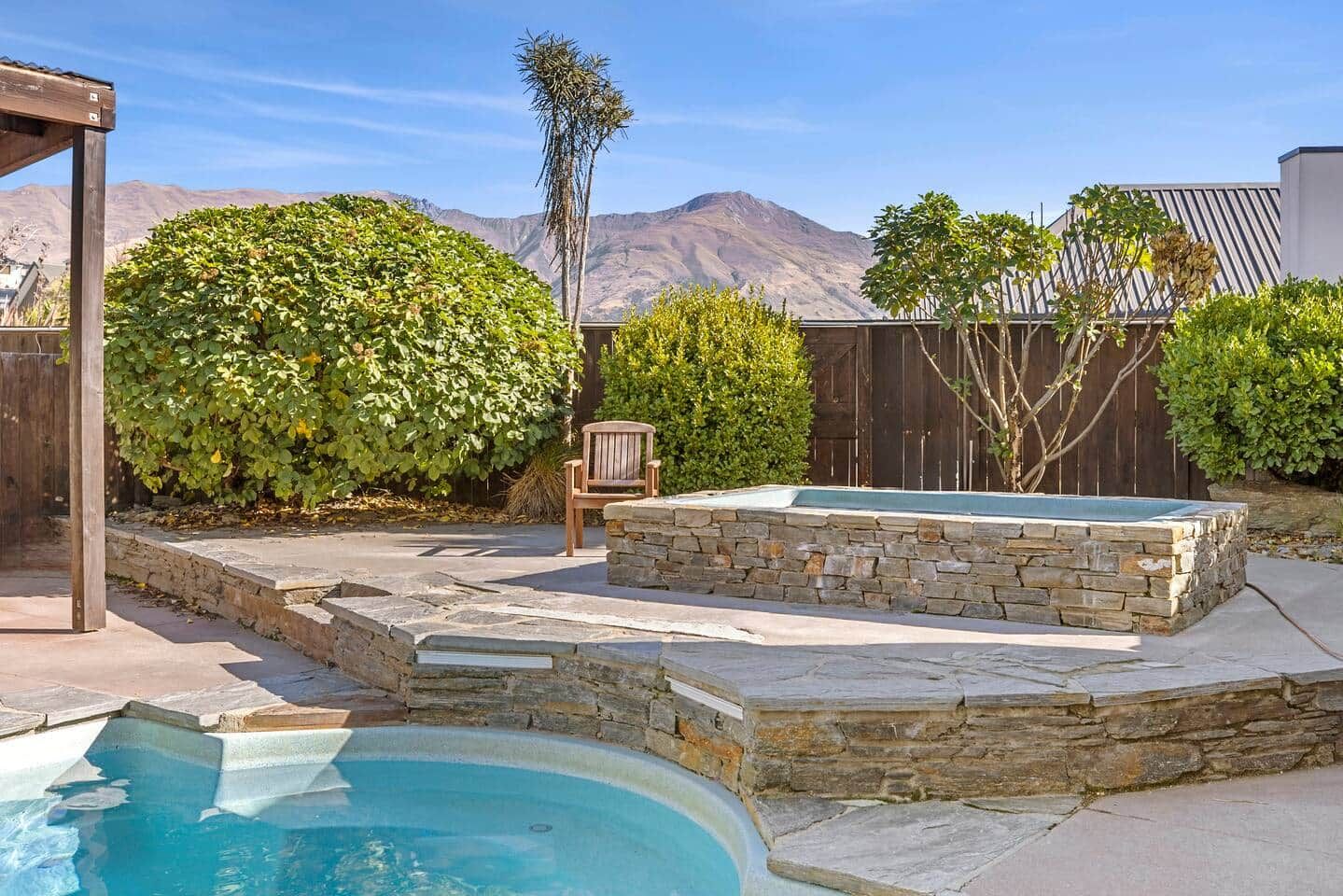 A backyard with a swimming pool, stone hot tub, wooden deck, and a chair.