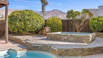 A backyard with a swimming pool, stone hot tub, wooden deck, and a chair.