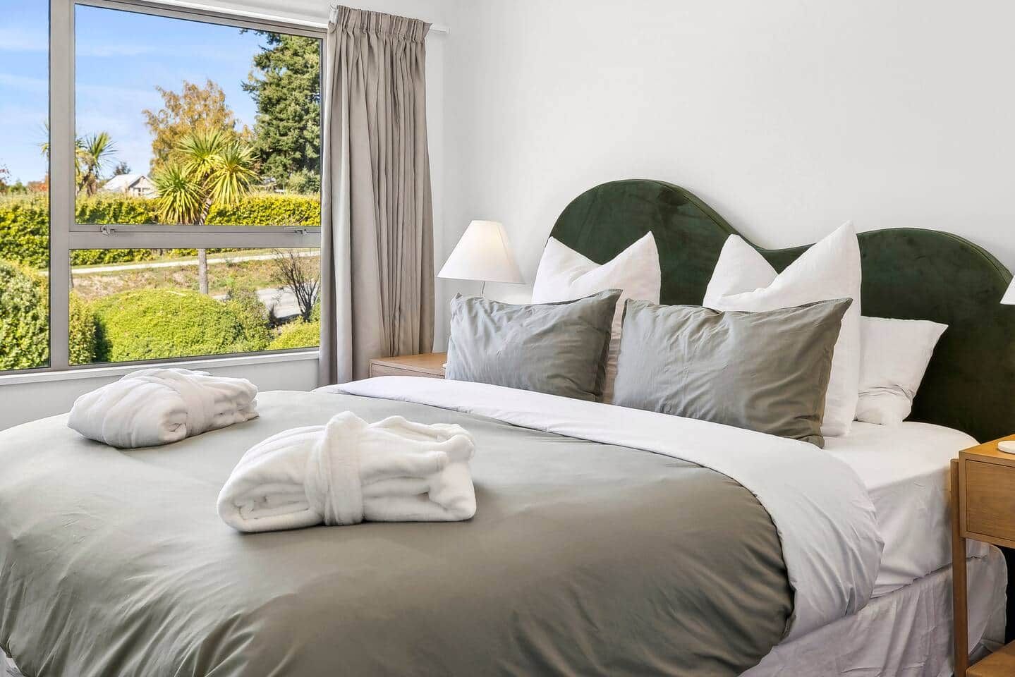 A neatly made bed with a headboard, a bedside lamp, and a view of greenery outside.