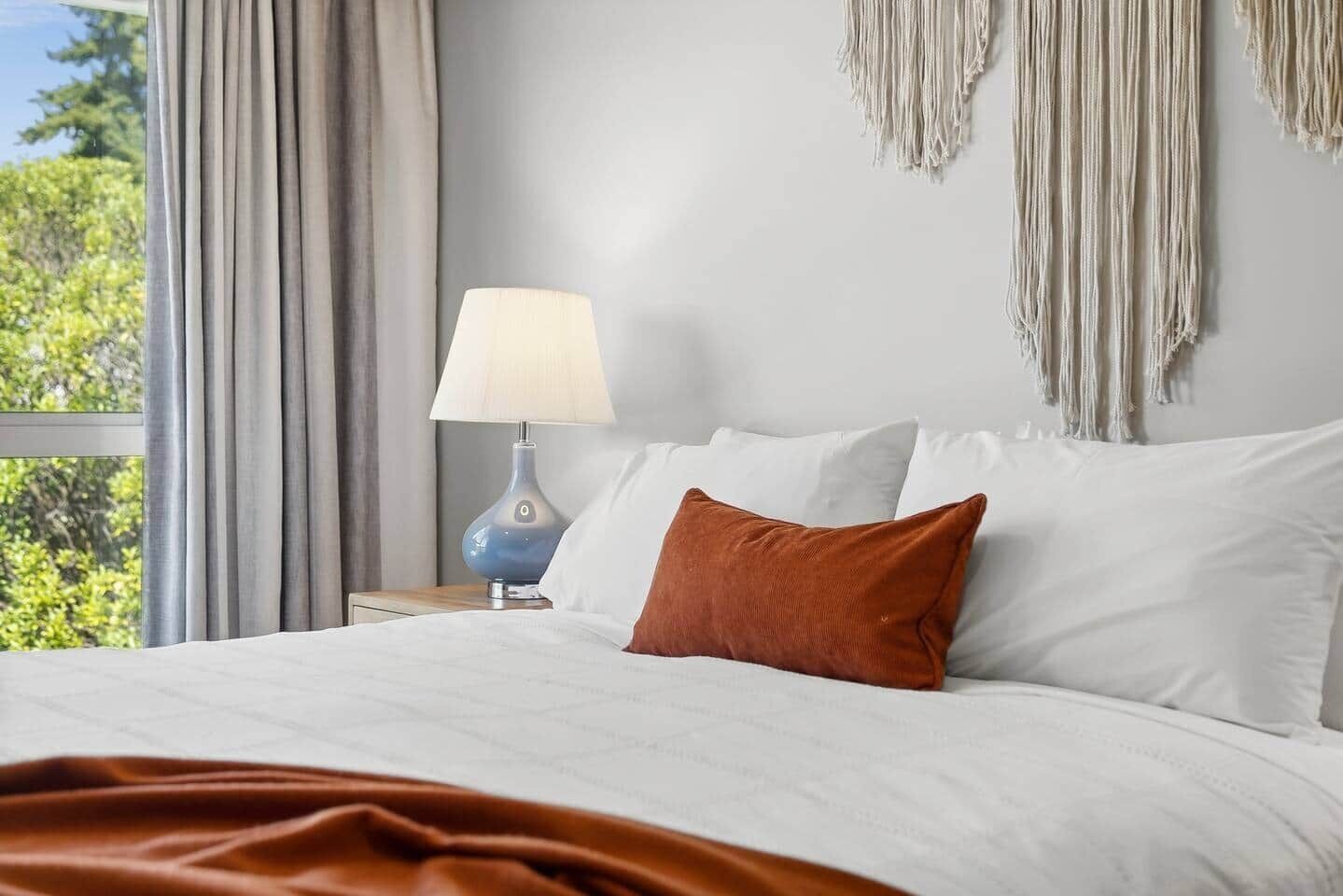 A neatly made bed with white and brown pillows, a blue lamp on a bedside table, and a view of greenery through a window.