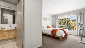A bedroom with a bed, a window with curtains, a standing fan, and a bathroom with a sink and mirror. - Mountain Four-Guest with Views (Wanaka)