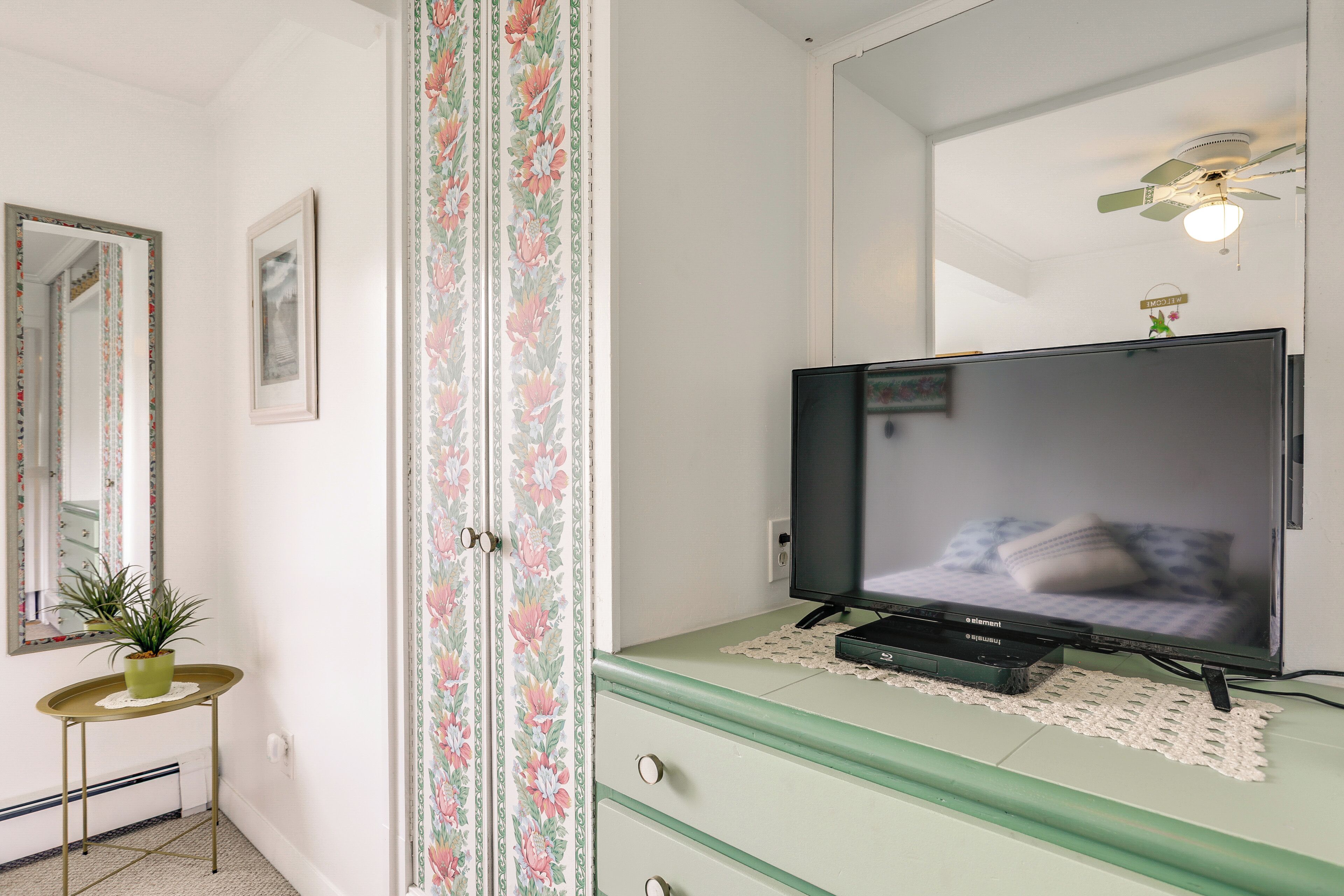 Une pièce avec une télévision posée sur une commode, un miroir au mur et un ventilateur de plafond.
