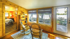 Ein Interieur einer Holzhütte mit einem Bett, Schaukelstühlen, einem Bücherregal und großen Fenstern mit Blick ins Freie.
