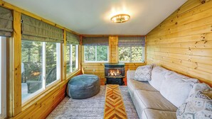 Ein gemütliches Blockhaus-Interieur mit Kamin, einer Couch, einem Sitzsack und großen Fenstern mit Blick ins Freie.