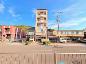 A multi-story building with balconies, a parking area, and a hilly background. - All rooms have ocean views | 3rd floor guest rooms / Koba City Fukui (Koba City)