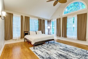 Ein Schlafzimmer mit Bett, Deckenventilator, großem Fenster mit Vorhängen und Holzboden.