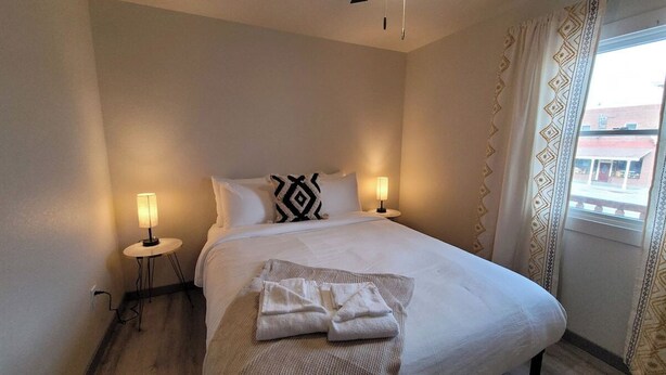 A hotel room with a bed, two bedside lamps, a window with curtains, and a small table with a lamp. - Jackalope Loft in Downtown Custer #2 (Custer)