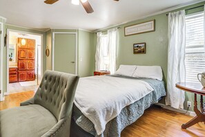 Ein Schlafzimmer mit einem Bett, einem Sessel, einem Nachttisch, einer Lampe und einem Fenster mit Vorhängen.