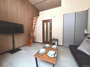 Ein Wohnzimmer mit Fernseher, einem Holztisch für vier Personen, einer Couch, einer Leiter und einer Tür.