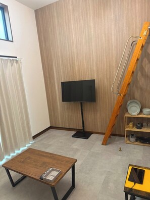 Ein Zimmer mit Holzboden, einem Fernseher auf einem Schrank, einem kleinen Tisch mit Tablett, einem Regal mit Geschirr und einer an der Wand angelehnten Leiter.