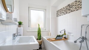 A modern bathroom with a white sink, bathtub, and shower. There is a window with a view of the outside, a potted plant, and a wooden shelf with toiletries. - Gemütliches Apartment in Dachau (Freilassing)