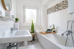 A modern bathroom with a white sink, bathtub, and shower. There is a window with a view of the outside, a potted plant, and a wooden shelf with toiletries.