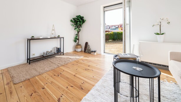 A modern living room with a wooden floor, a black console table, a white sofa, and a large window with a view of buildings and greenery. - Gemütliches Apartment in Dachau (Freilassing)