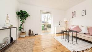 A modern living room with a wooden floor, a white sofa, a coffee table, a side table, a potted plant, and a bookshelf. - Gemütliches Apartment in Dachau (Freilassing)