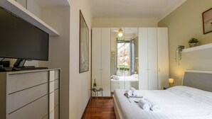 Ein Hotelzimmer mit Bett, Fernseher, Kleiderschrank, einem kleinen Tisch mit Flasche und Gläsern sowie einem Spiegel, der einen Balkon reflektiert.