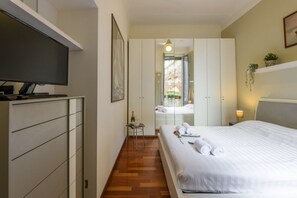 Ein Hotelzimmer mit Bett, Fernseher, Kleiderschrank, einem kleinen Tisch mit Flasche und Gläsern sowie einem Spiegel, der einen Balkon reflektiert.