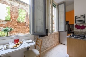 A kitchen with a brick wall, a window with open blinds, a table with a tray of food, and a chair. - 81 Garibaldi – Brera Base   (Milan)