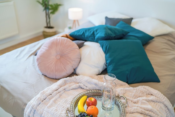 Un lit bien fait, agrémenté de plusieurs coussins, et une table d’appoint avec des fruits et un verre d’eau.