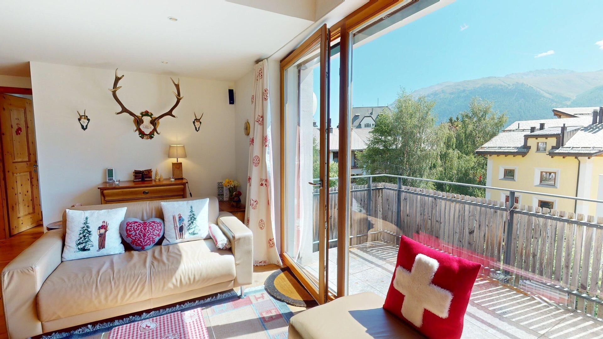 Ein gemütliches Wohnzimmer mit einer Couch, dekorativen Kissen, einem Holztisch und Blick auf einen Balkon mit Holzlattenzaun und Gebäuden im Hintergrund.