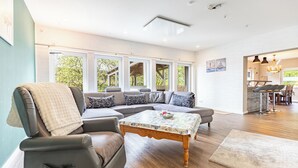 Ein modernes Wohnzimmer mit einem Ecksofa, einem Holztisch und großen Fenstern mit Blick ins Freie.