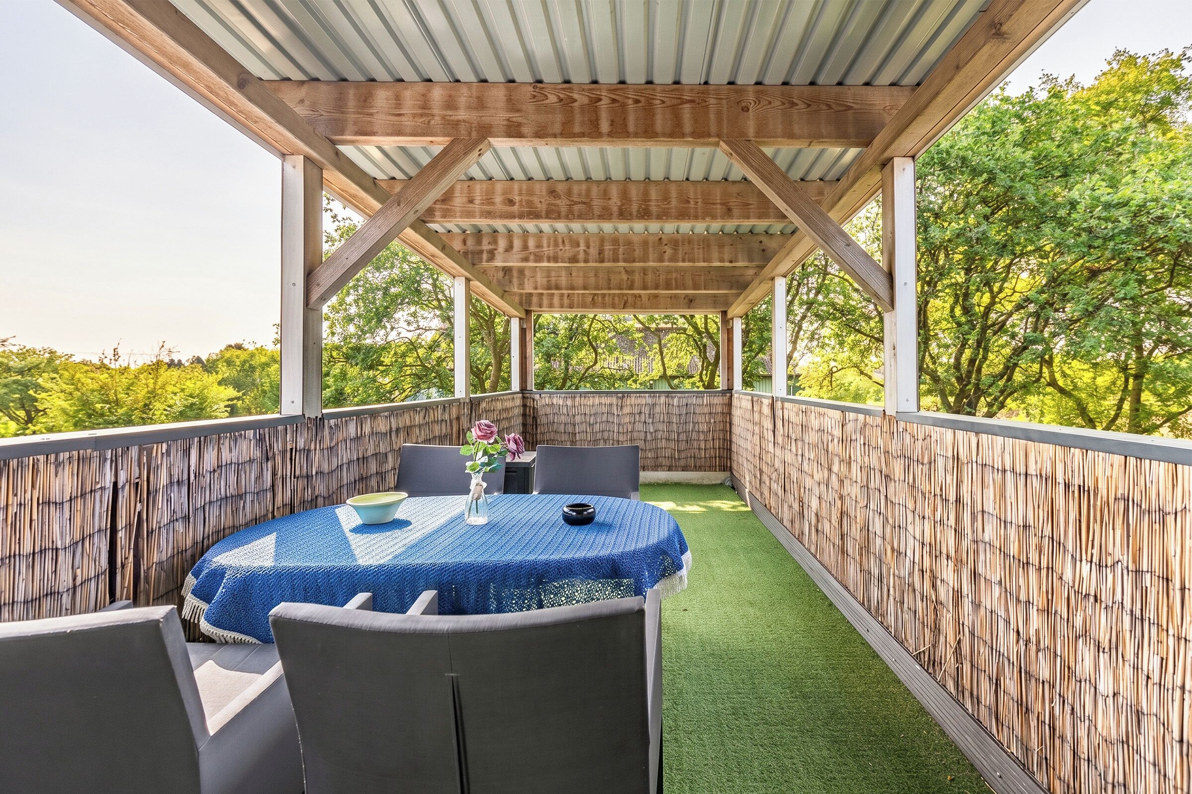 Eine überdachte Terrasse im Freien mit Tisch und Stühlen, umgeben von einem Bambuszaun und grünem Kunstgrün.