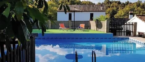 Una piscina con dos sillas reclinables, un pequeño edificio con techo de tejas y una cerca de madera.
