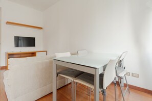 Ein modernes Wohnzimmer mit Esstisch aus Glas, weißen Stühlen, einer Couch und einem flachbildfernseher an der Wand.