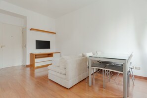Ein modernes Wohnzimmer mit einer Couch, einem Esstisch mit Glasplatte, einem an der Wand montierten Fernseher und einem Holzregal.