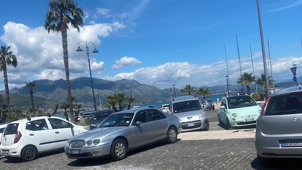 Un estacionamiento con coches, palmeras y montañas al fondo.