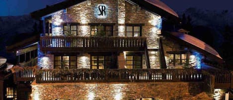 A multi-story stone building with a prominent 'SR' logo, balconies, and a reflective water feature in front.