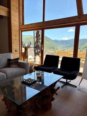 Ein modernes Wohnzimmer mit einem Glastisch, zwei schwarzen Sesseln, einem grauen Sofa und einem großen Fenster mit Bergblick.