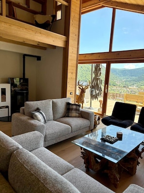 Ein geräumiger Wohnbereich mit hoher Decke, einem Glastisch und Blick auf eine gebirgige Landschaft.