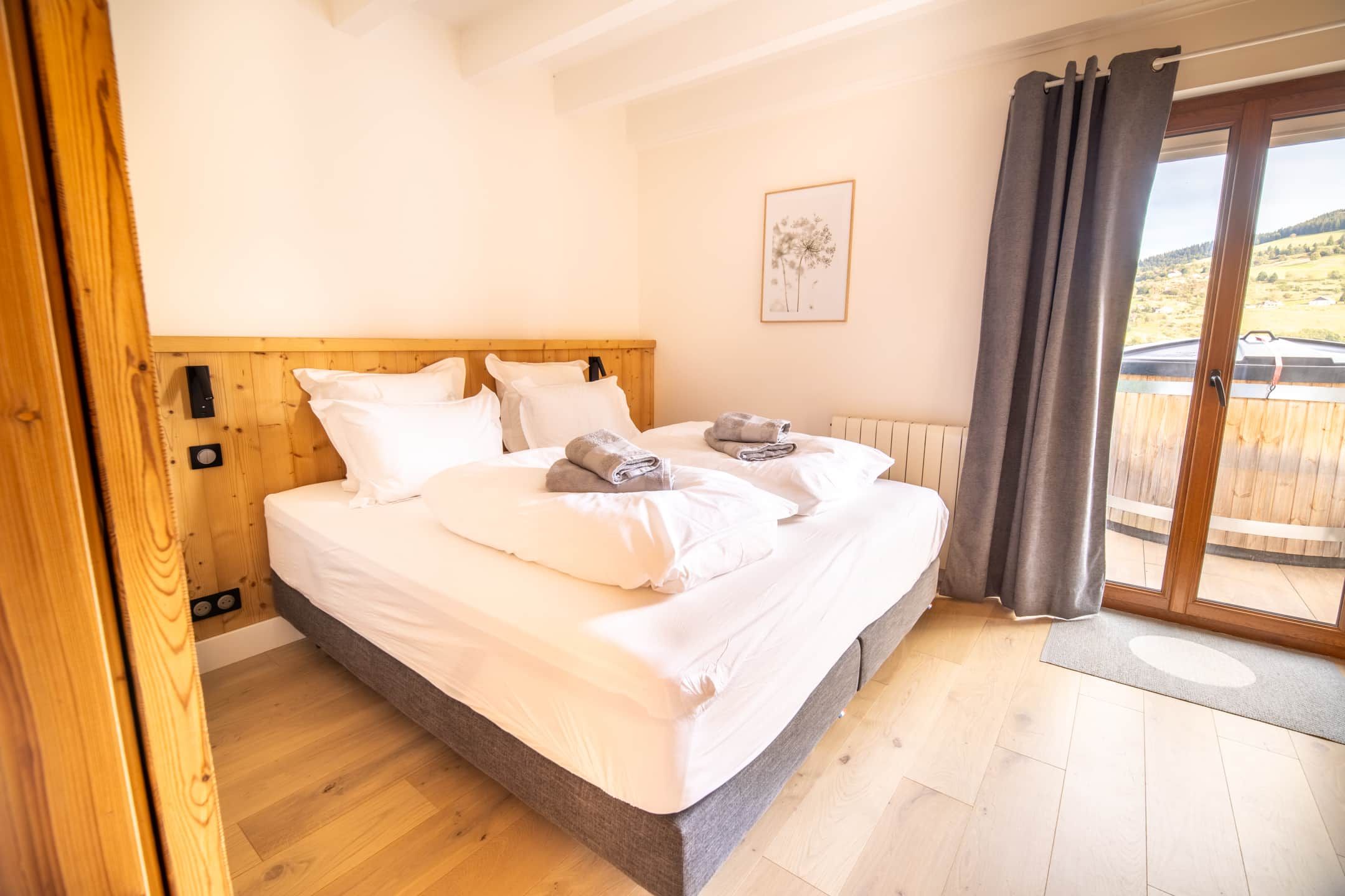 Ein Schlafzimmer mit einem hölzernen Kopfteil, einem großen Bett mit weißer Bettwäsche, einer grauen Tagesdecke und Blick auf eine Holzterrasse durch eine Glastür.
