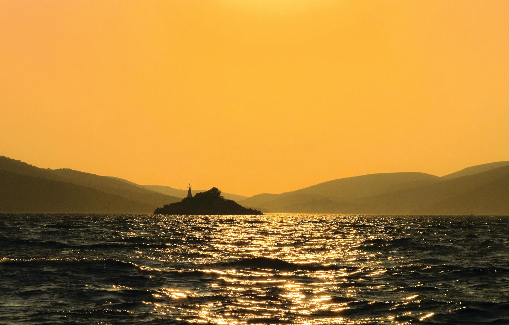 Silhouet van een vuurtoren tegen een ondergaande zon, weerspiegeld in het water, met heuvels op de achtergrond.