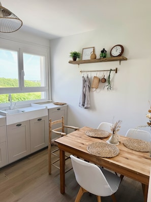 Eine Küche mit weißen Schränken, einem Holzespeisetisch mit gewebten Tischsets und einem Fenster mit Blick ins Grüne.