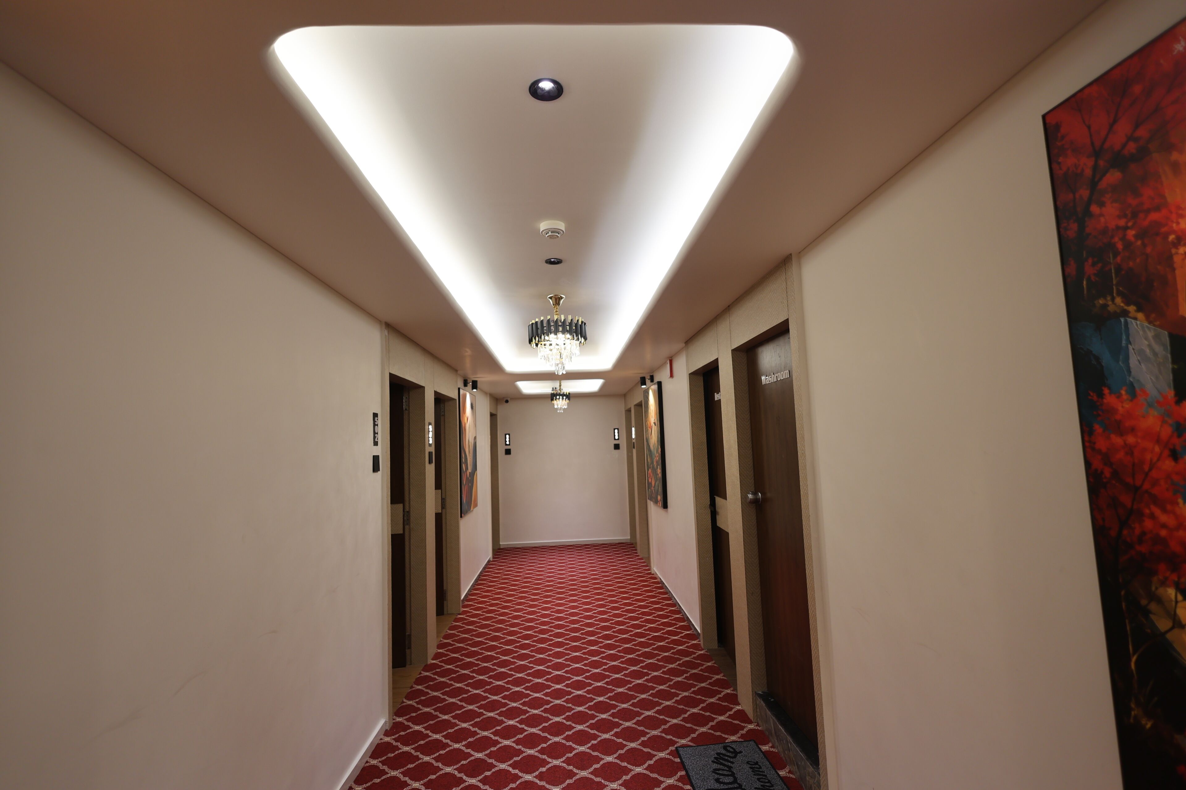 Un pasillo de hotel con alfombra roja estampada, una lámpara de araña y un cuadro en la pared derecha.