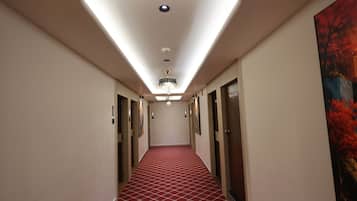 Un pasillo de hotel con alfombra roja estampada, una lámpara de araña y un cuadro en la pared derecha.