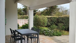Um alpendre coberto com uma mesa e cadeiras, rodeado por um jardim.