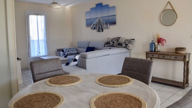 Uma sala de estar com um sofá cinzento, duas poltronas, uma mesa de centro com jogos americanos trançados, uma mesa de jantar, um quadro na parede e uma pequena mesa lateral de madeira com objetos decorativos.