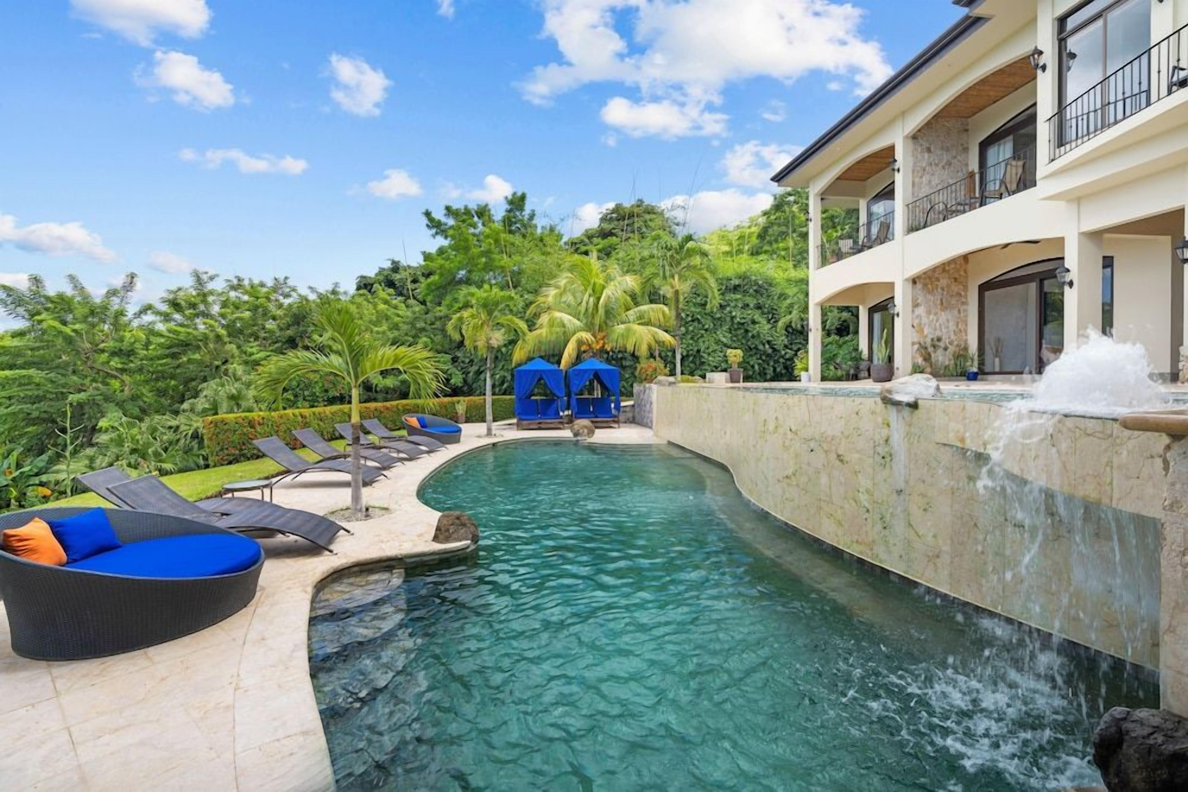 A swimming pool with lounge chairs and a waterfall feature, surrounded by lush greenery and a multi-story building.