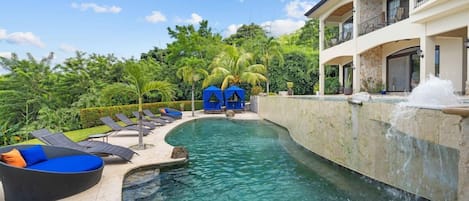 A swimming pool with lounge chairs and a waterfall feature, surrounded by lush greenery and a multi-story building.