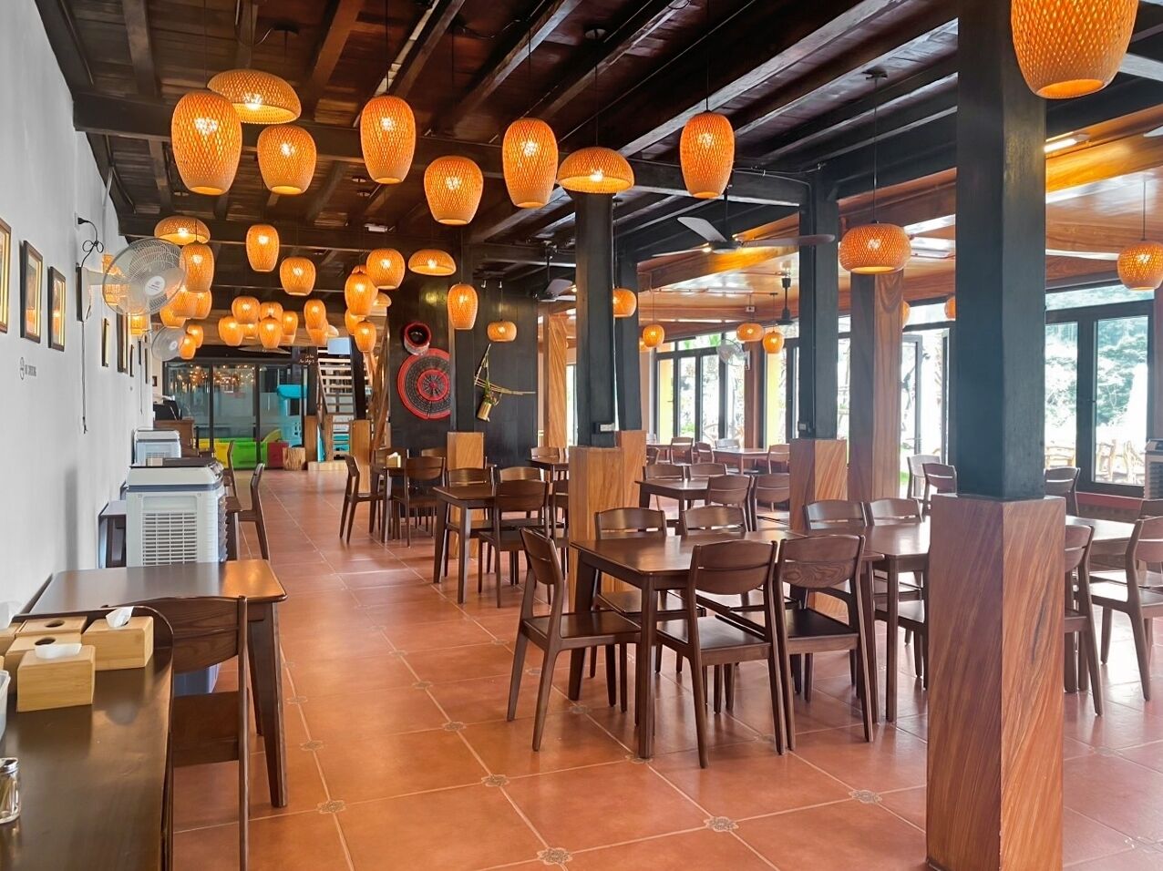 A restaurant with wooden tables and chairs, hanging lanterns, and a large window in the background.