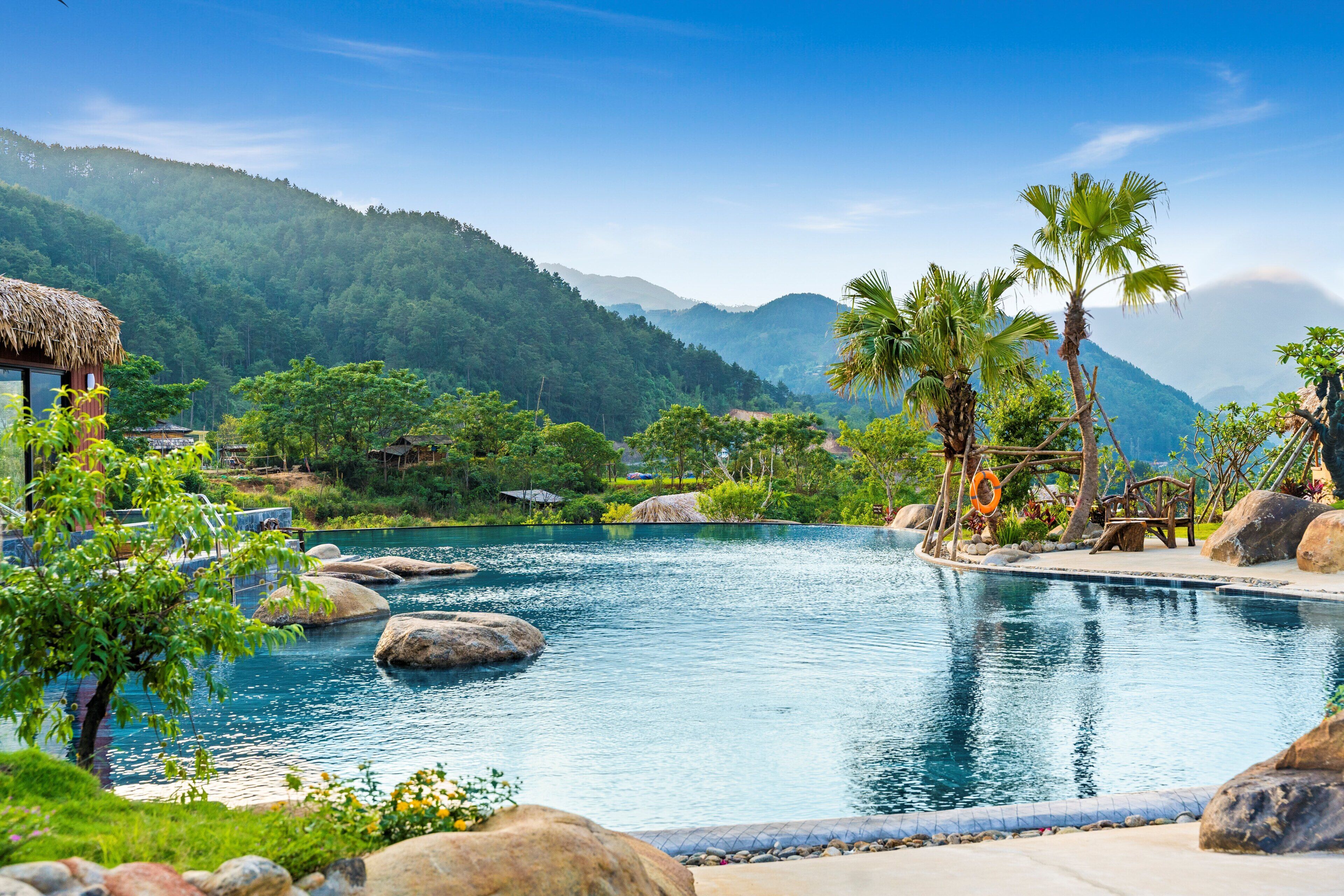 A pool with rocks and palm trees, surrounded by greenery and mountains.