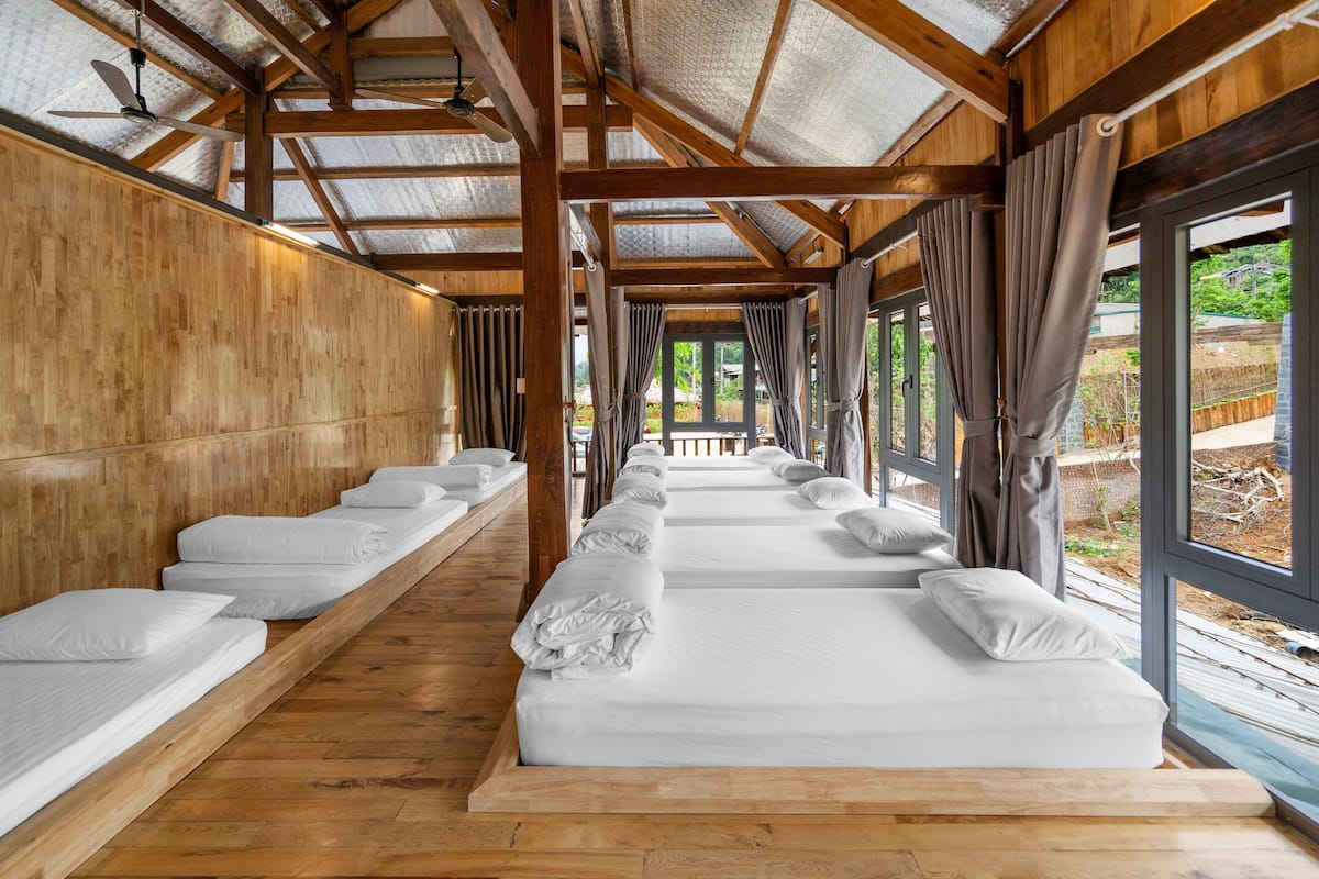 A row of single beds with wooden headboards, a ceiling fan, and large windows with curtains.