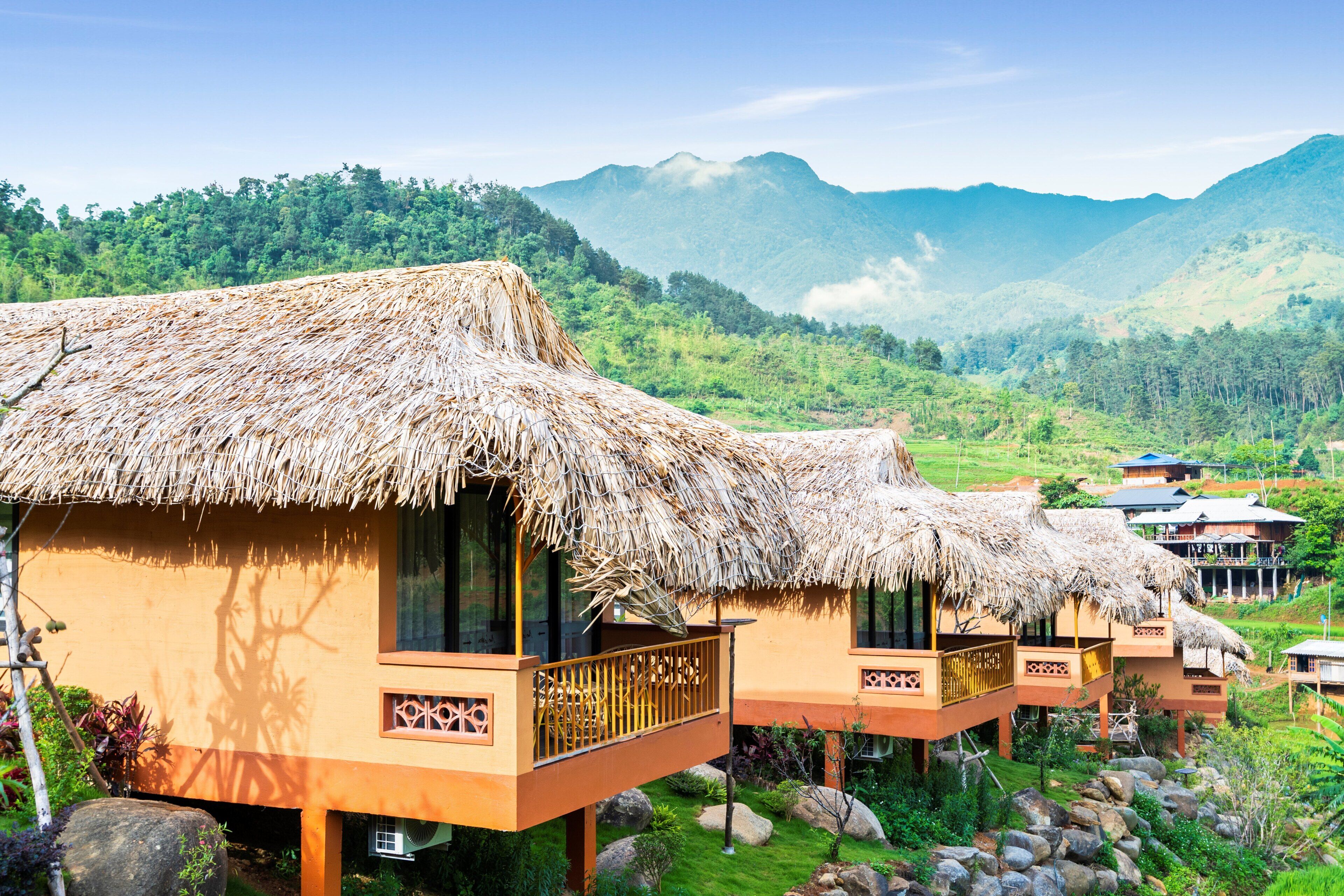 Thatched-roof cottages with balconies, set in a lush valley with mountains in the background.