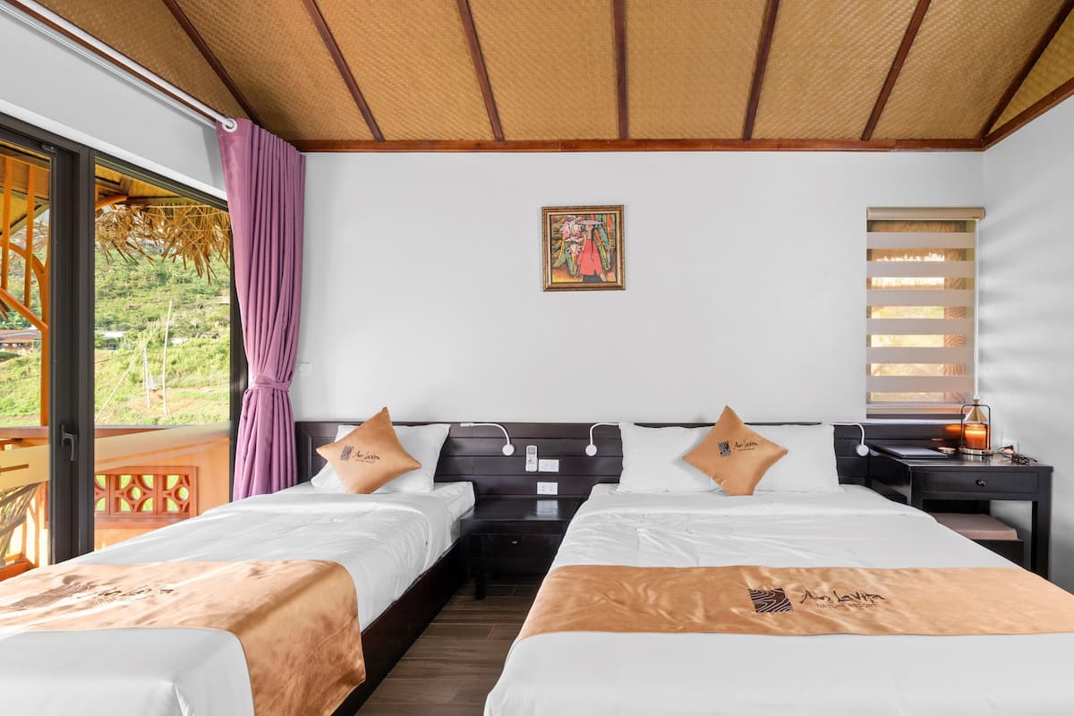 A hotel room with two beds, a wooden headboard, a nightstand, a window with blinds, and a view of greenery outside.